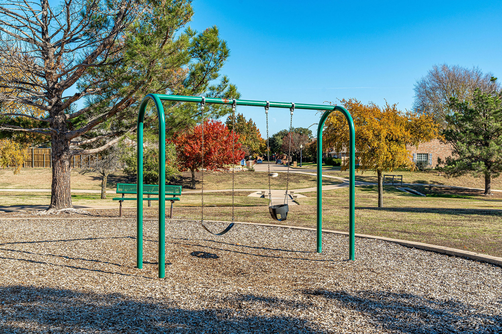 A green swing set with two swings stands in a park surrounded by trees, benches, and a wood chip ground covering under a clear blue sky.