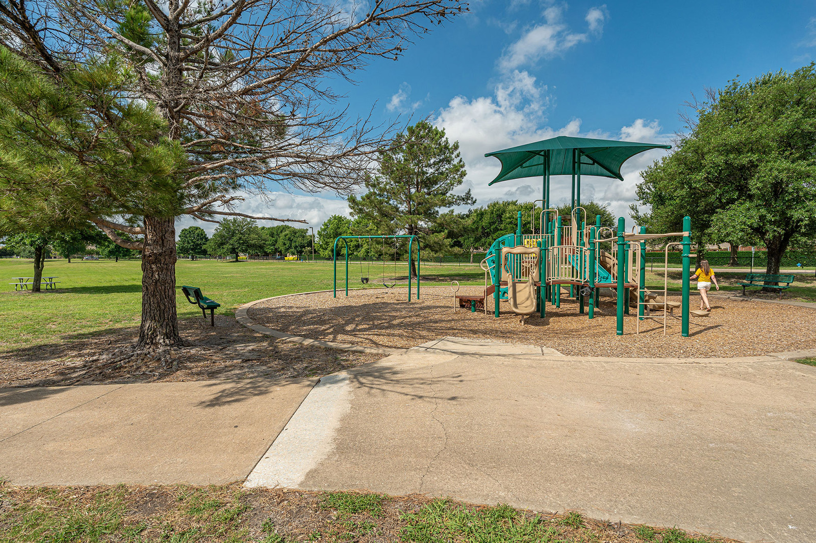 A playground with slides, swings, and climbing structures sits on a mulch surface in a park with green grass, trees, benches, and a blue sky with scattered clouds.