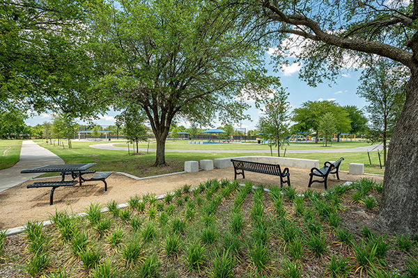 A park with benches, picnic tables, a circular walking path, green grass, trees, and landscaped plants under a blue sky.