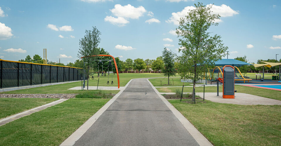 A paved path runs through a park with green grass, young trees, a playground area, and a black chain-link fence under a partly cloudy sky.