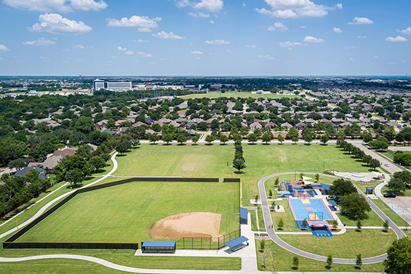 Aerial view of a public park with a baseball field, walking paths, a playground, and residential houses in the background under a partly cloudy sky.