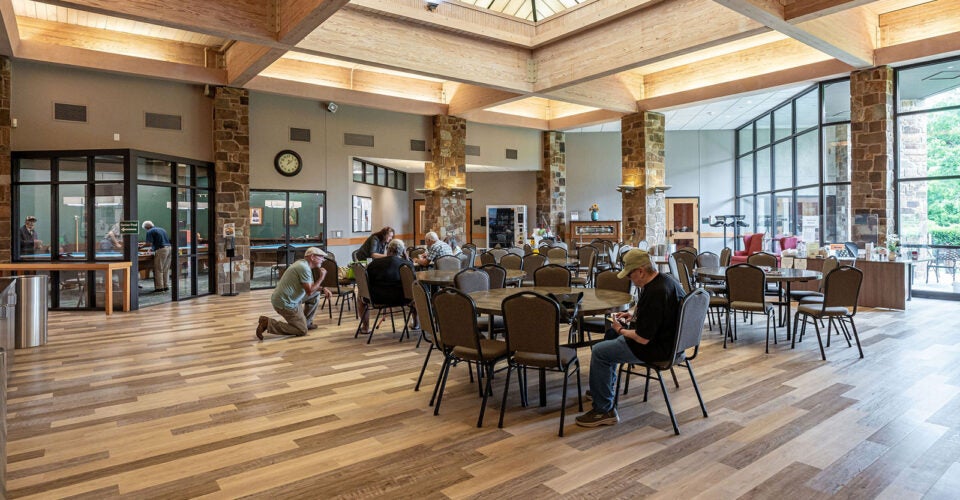 People sit, read, and talk at tables in a spacious community center with large windows, wooden beams, and a vaulted ceiling with natural light.