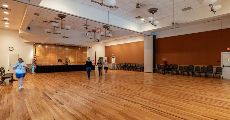 A spacious indoor hall with a wooden floor, stage at the back, rows of chairs along the walls, and several people walking or standing in the room.
