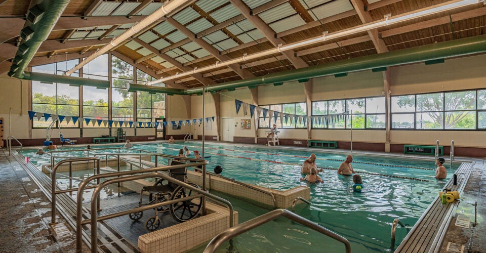 Indoor pool with several people swimming and exercising; a wheelchair and pool lift are visible in the foreground. Large windows let in natural light.