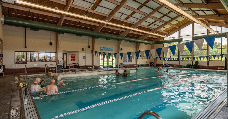 Indoor swimming pool with several people in the water and others seated at the edge; large windows and triangular flags decorate the space.