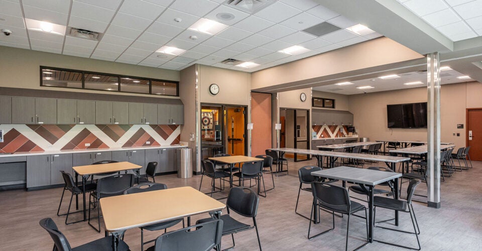 Modern cafeteria with black chairs and light wood tables, wall-mounted clocks, television, and a kitchenette area with cabinets and countertops. Room is empty and well-lit.