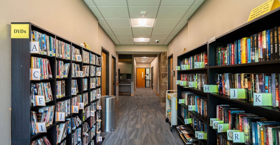 Library hallway with bookshelves on both sides, alphabetized labels on shelves, DVDs on the left, books on the right, and a recycling bin in the center.