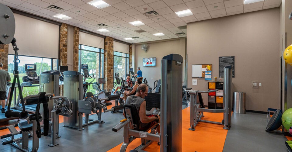 People use various exercise machines in a well-lit gym with large windows, orange and gray flooring, and fitness equipment arranged throughout the room.