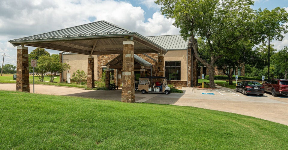 A stone and metal building entrance with a covered drive, parked golf cart, cars, and surrounding grassy area under a partly cloudy sky.