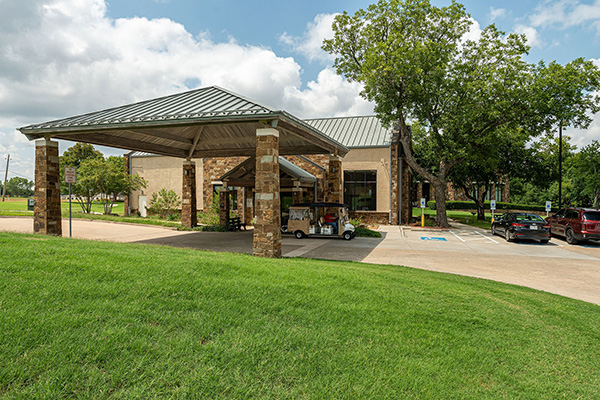 Single-story building with a covered entrance, stone accents, and a golf cart parked outside; surrounded by grass, trees, and a parking lot with several cars.