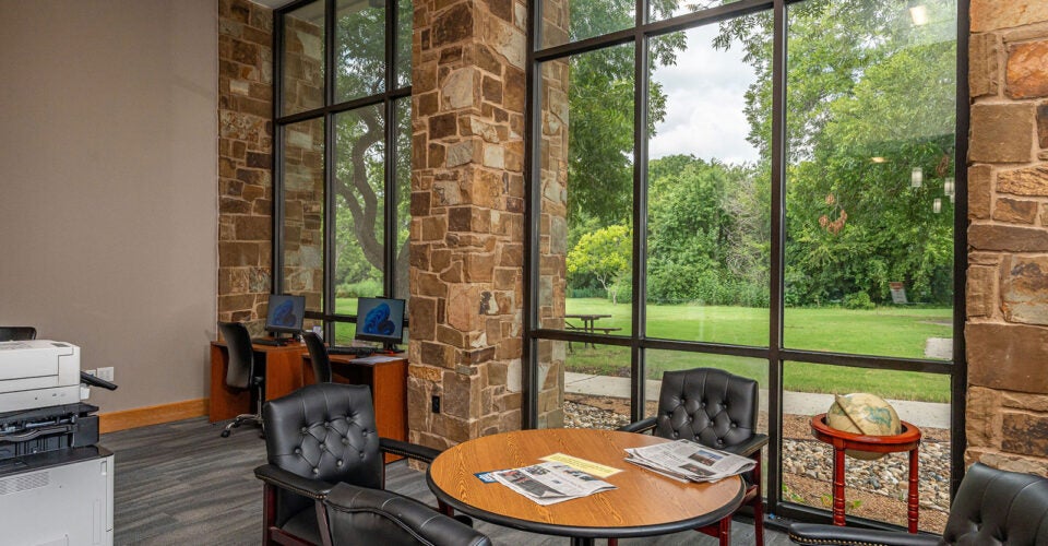 Office lounge area with leather chairs, a round table with newspapers, computer desks, a printer, and large windows overlooking a green outdoor landscape.