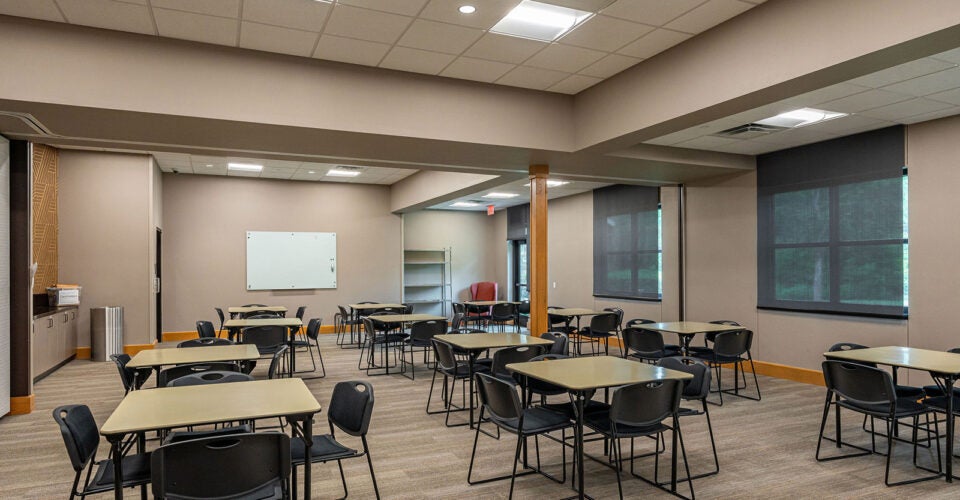 A classroom with several tables and chairs arranged in small groups, a whiteboard on the wall, large windows, and neutral-colored walls and carpeted floor.