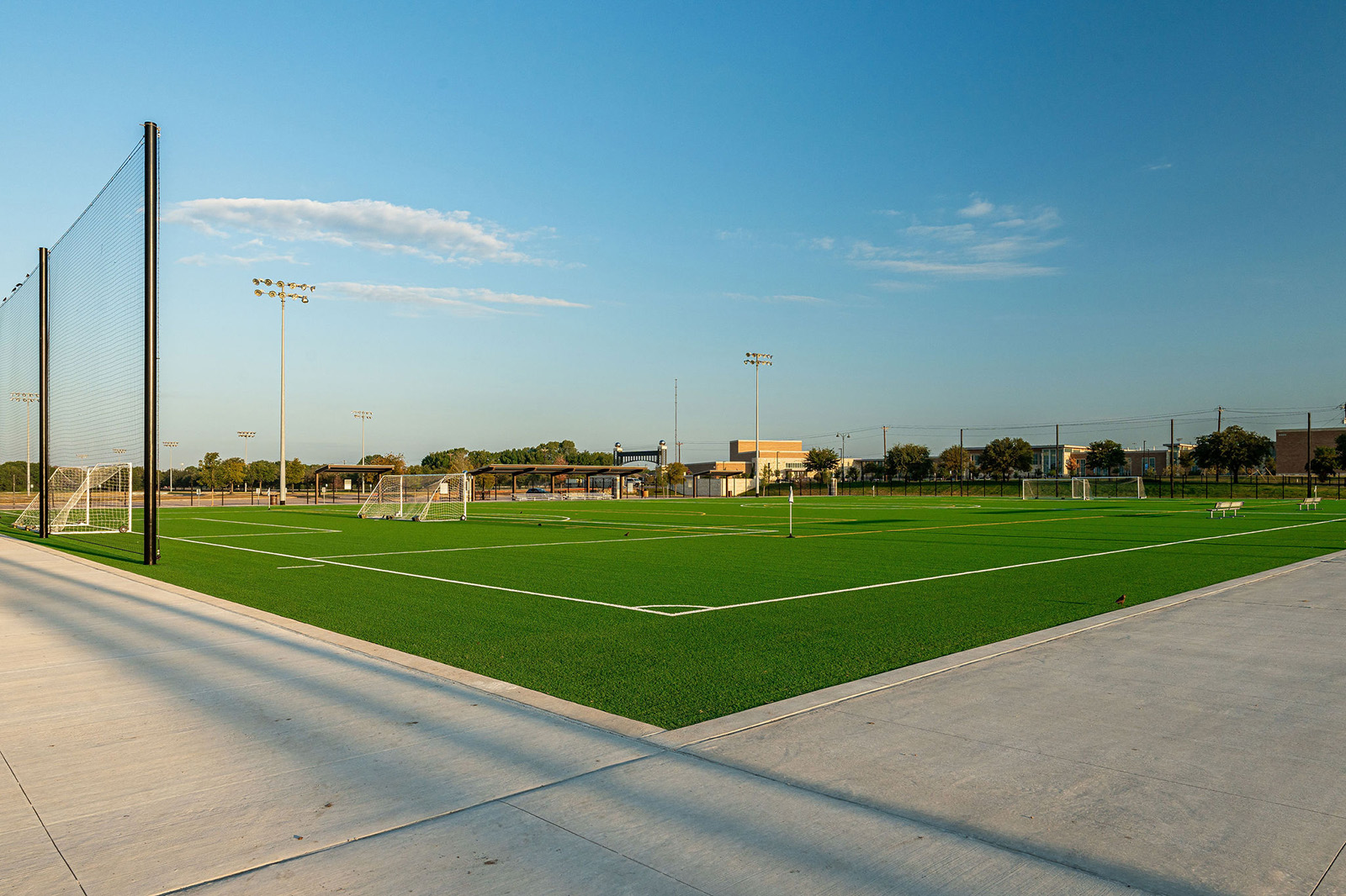 A large, empty artificial turf soccer field with multiple goals under clear blue sky, bordered by a concrete walkway and fencing.
