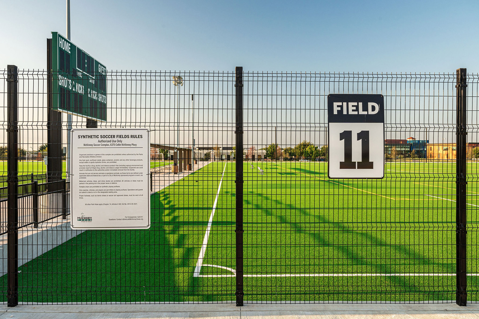 A fenced synthetic soccer field labeled "Field 11" with a posted sign of rules and a scoreboard visible in the background.