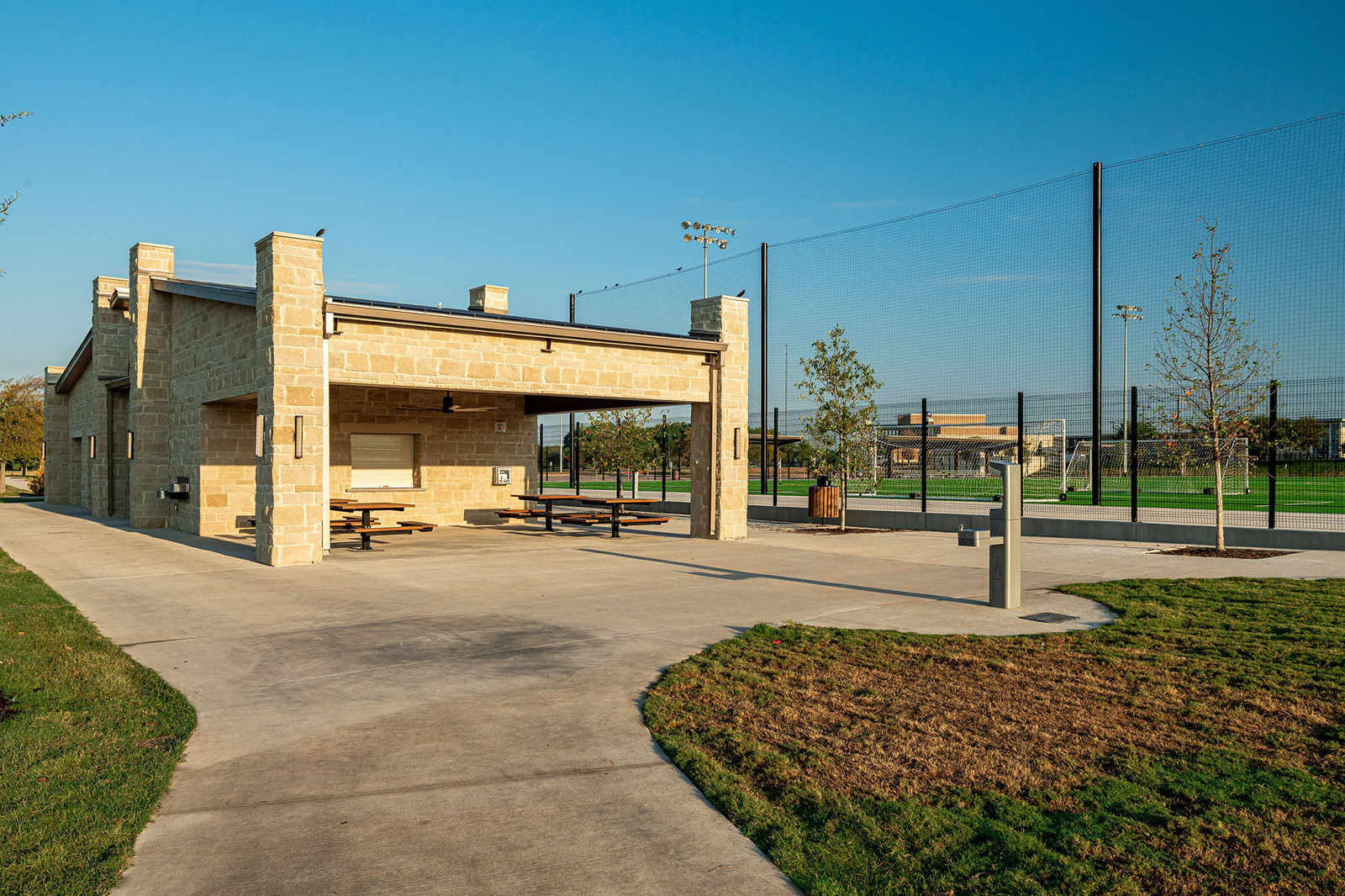 A stone pavilion with picnic tables is next to a fenced sports field under a clear sky in a park setting.