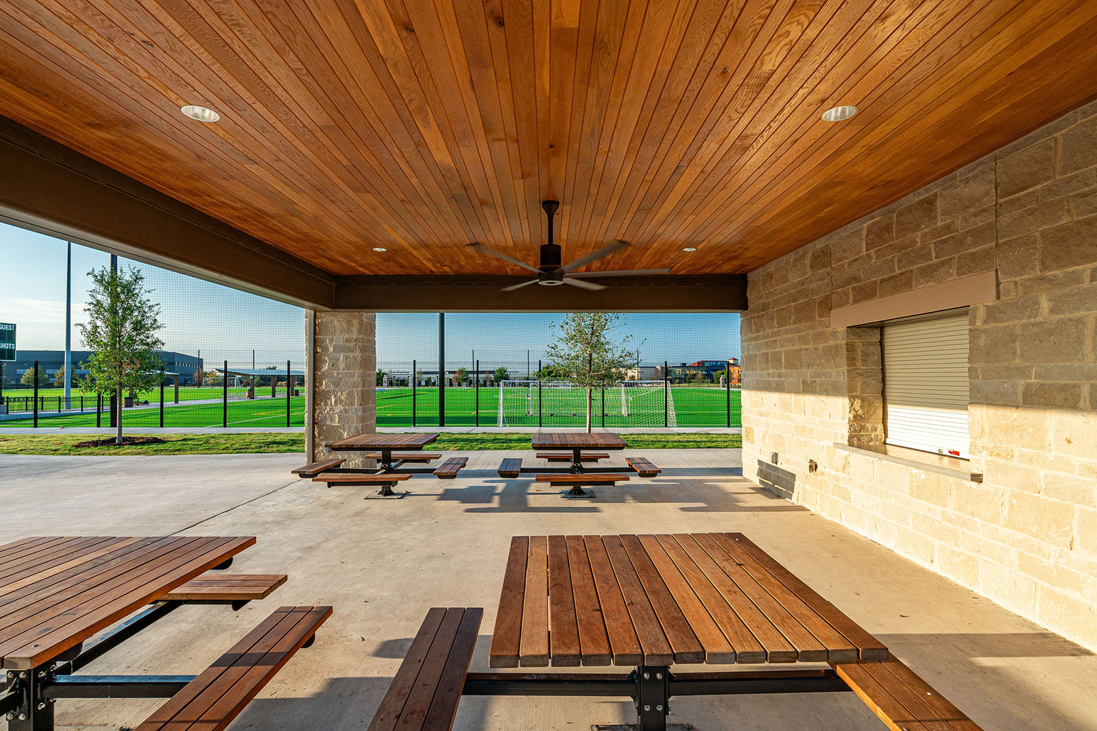 Covered outdoor pavilion with wooden picnic tables, a ceiling fan, and a serving window, overlooking a grassy sports field and fenced area on a sunny day.