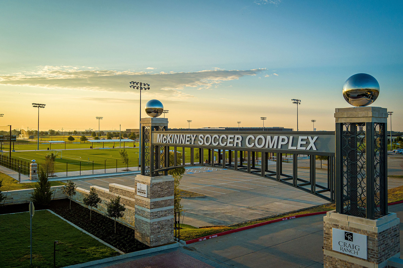 Entrance to McKinney Soccer Complex at Craig Ranch, featuring an archway and decorative pillars, with soccer fields and light poles visible in the background at sunrise.