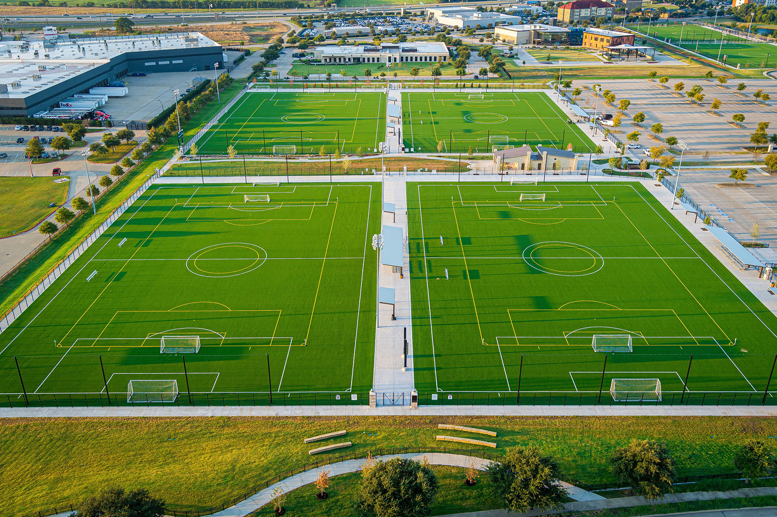 Aerial view of four artificial turf soccer fields with white markings, surrounded by parking lots and buildings in the background.