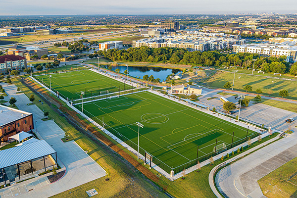 Aerial view of two artificial turf soccer fields surrounded by parking lots, buildings, a pond, and green spaces in an urban area.