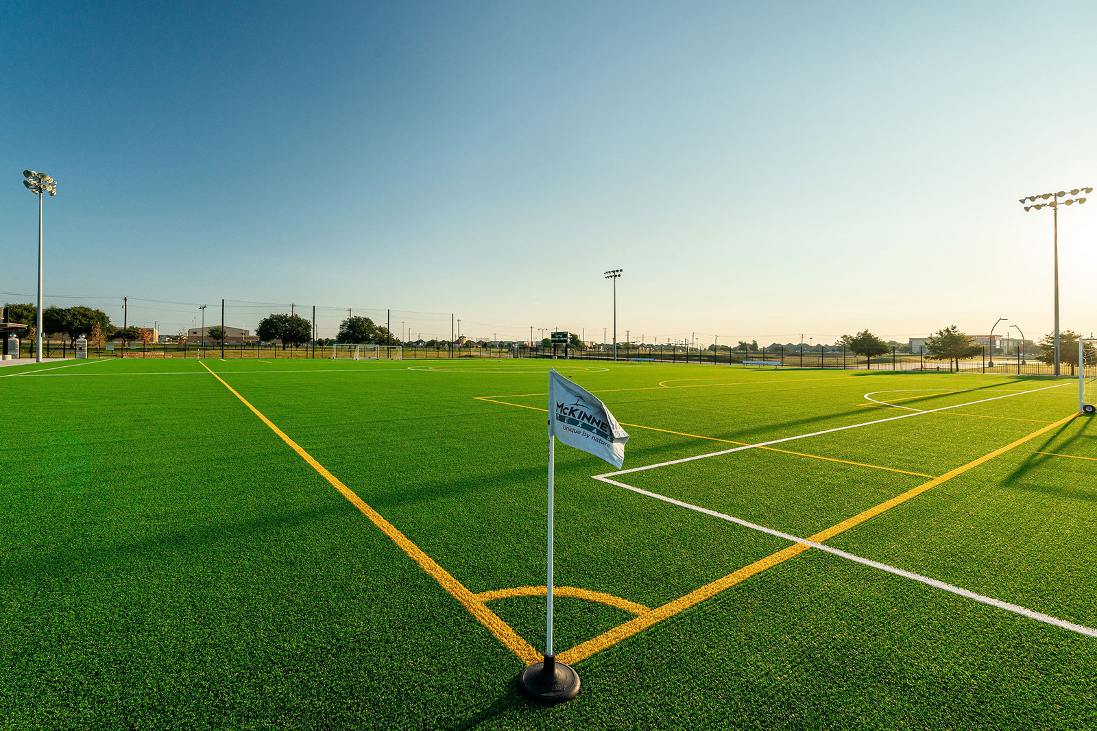 A well-maintained outdoor soccer field with bright green artificial turf, yellow and white field markings, and a corner flag, under a clear sky.