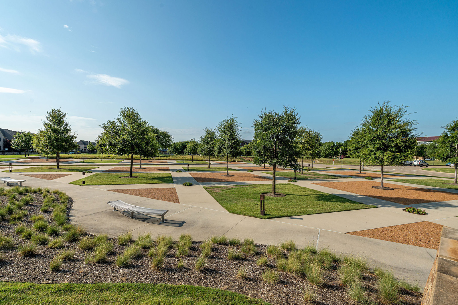 A park with neatly arranged trees, green grass patches, tan gravel squares, concrete walkways, benches, and manicured landscaping under a clear blue sky.