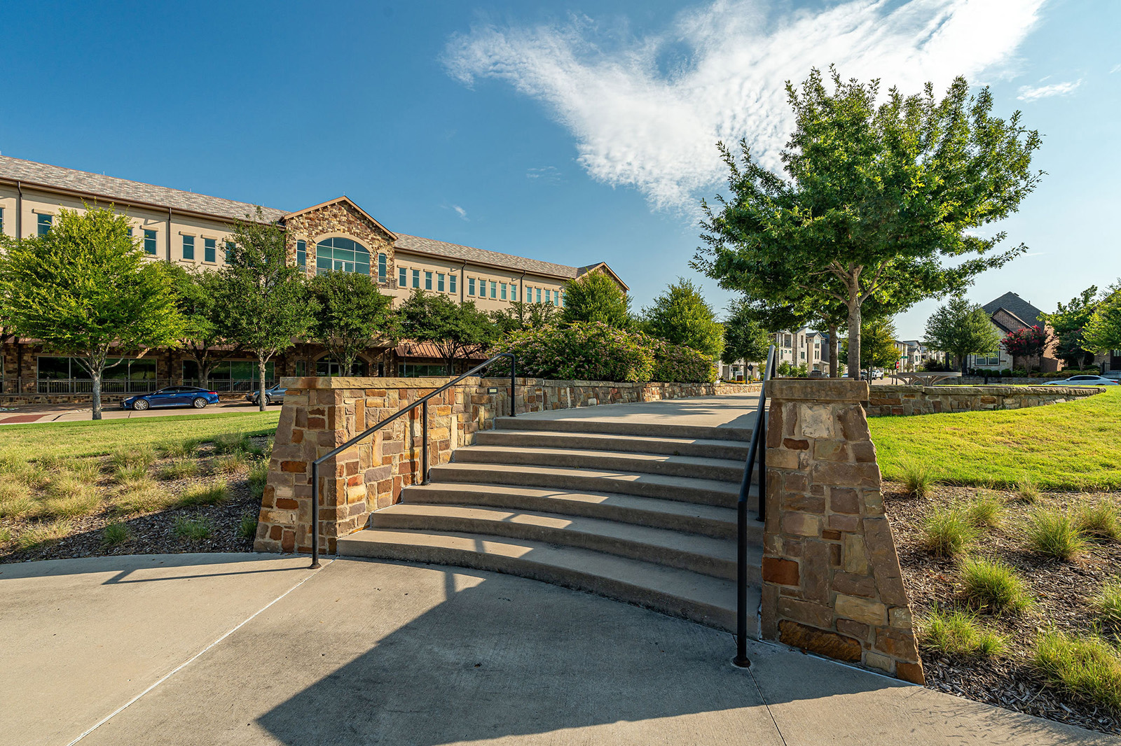Stone steps with metal railings lead up to a grassy area with trees and a large building in the background under a clear blue sky.