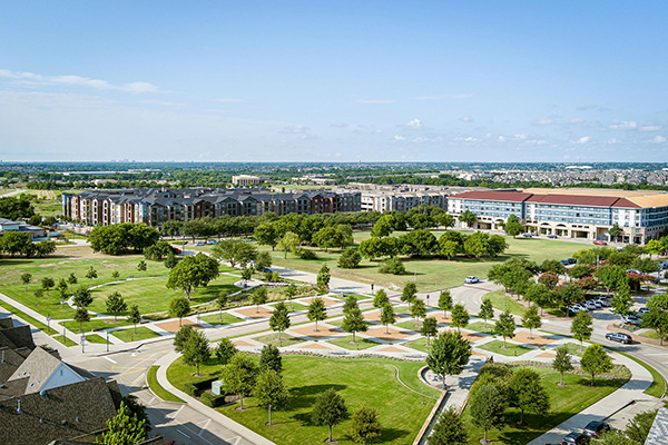Aerial view of a suburban area with apartment buildings, a large green park, pathways, and scattered trees under a clear blue sky.
