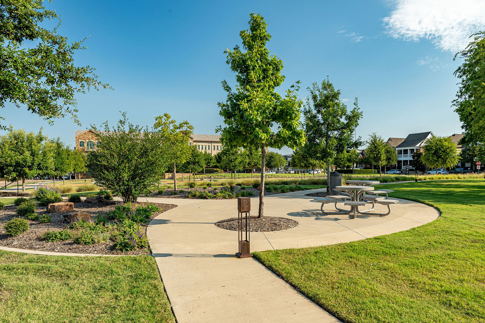 A paved walkway curves through a grassy park with trees, shrubs, a picnic table, and nearby buildings under a clear blue sky.