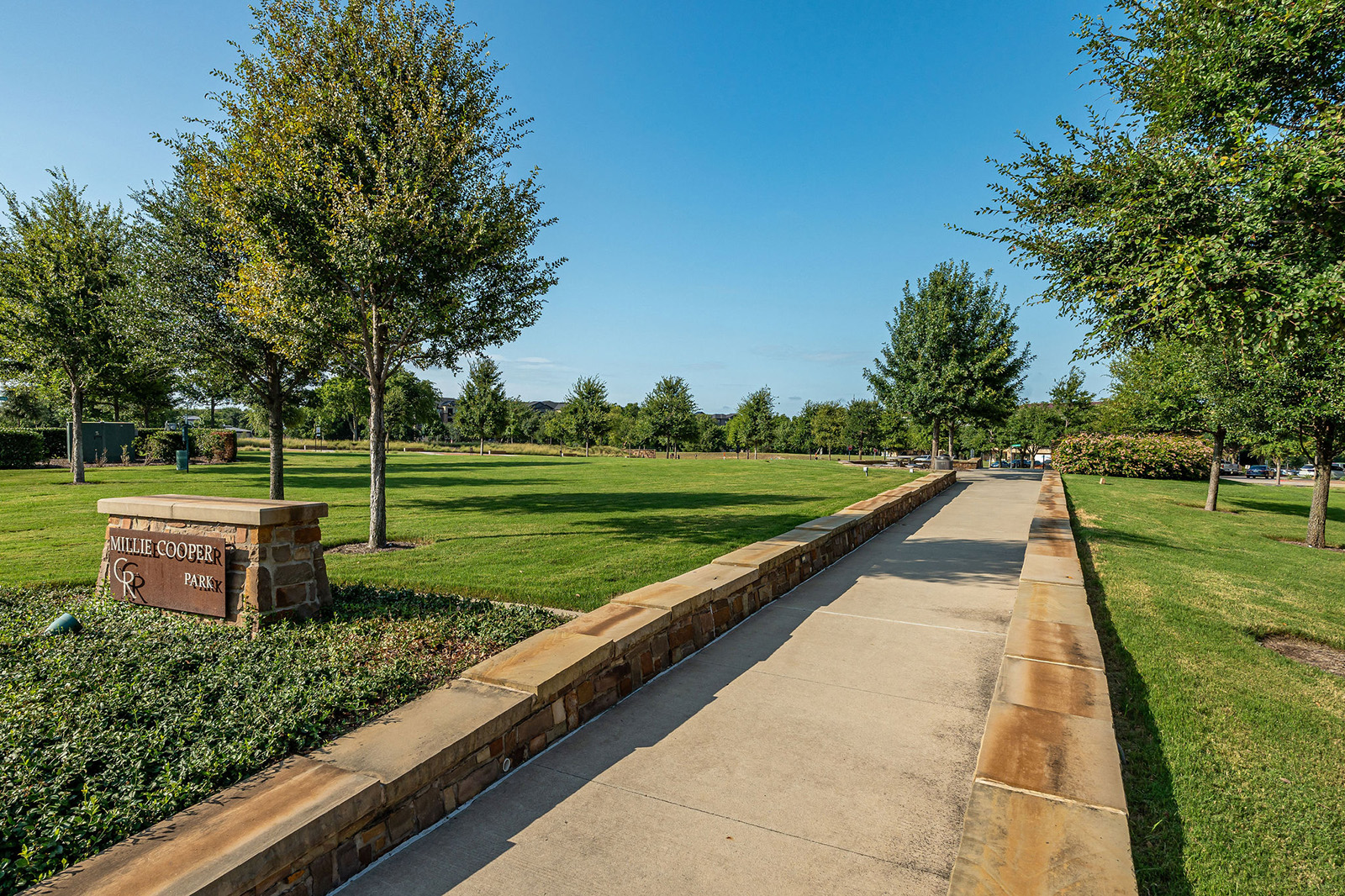 A paved walkway lined with stone borders runs through a grassy park with trees and a sign reading "Willie Cooper Park" on the left.