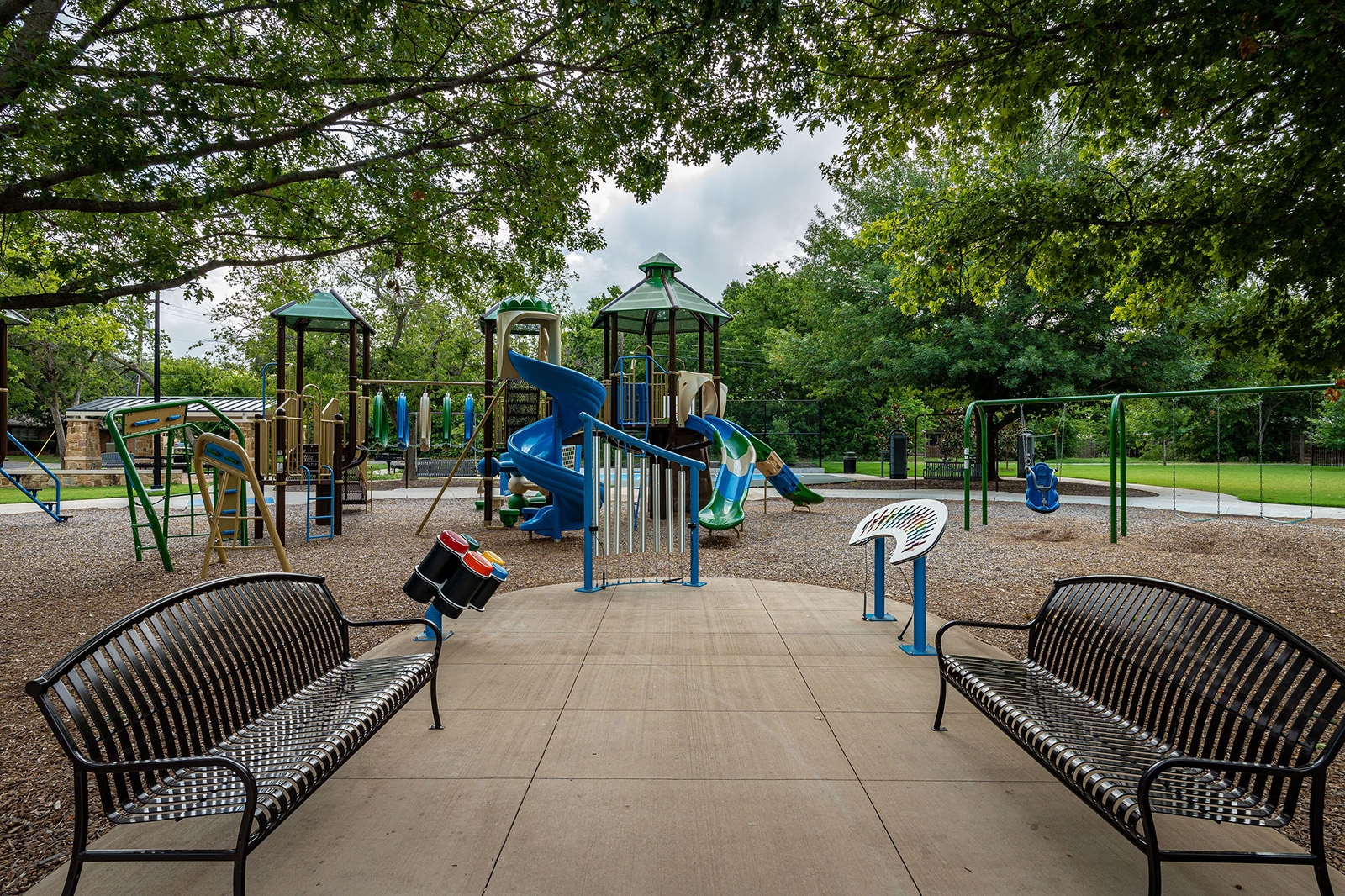 A playground with climbing structures and spiral slides is surrounded by trees, benches, and sensory play panels on a paved path.