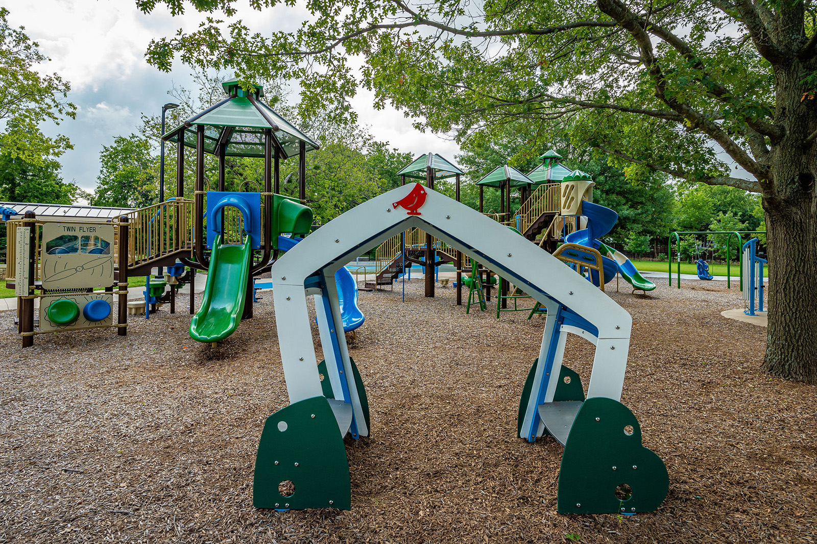 A playground with slides, climbing structures, and mulch ground covering, surrounded by trees under a cloudy sky.
