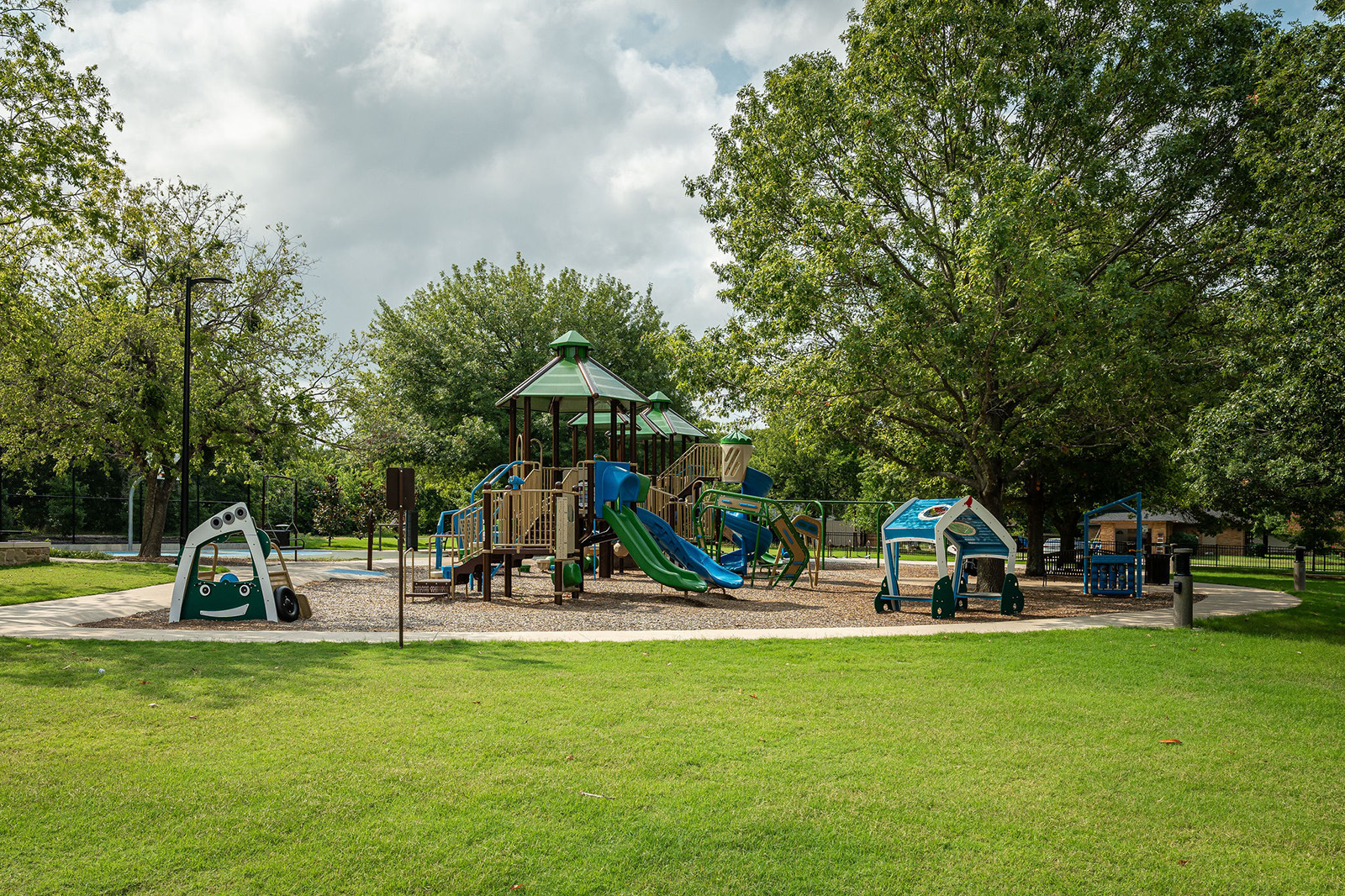A playground with slides, climbing structures, and toy vehicles is surrounded by trees and green grass under a partly cloudy sky.