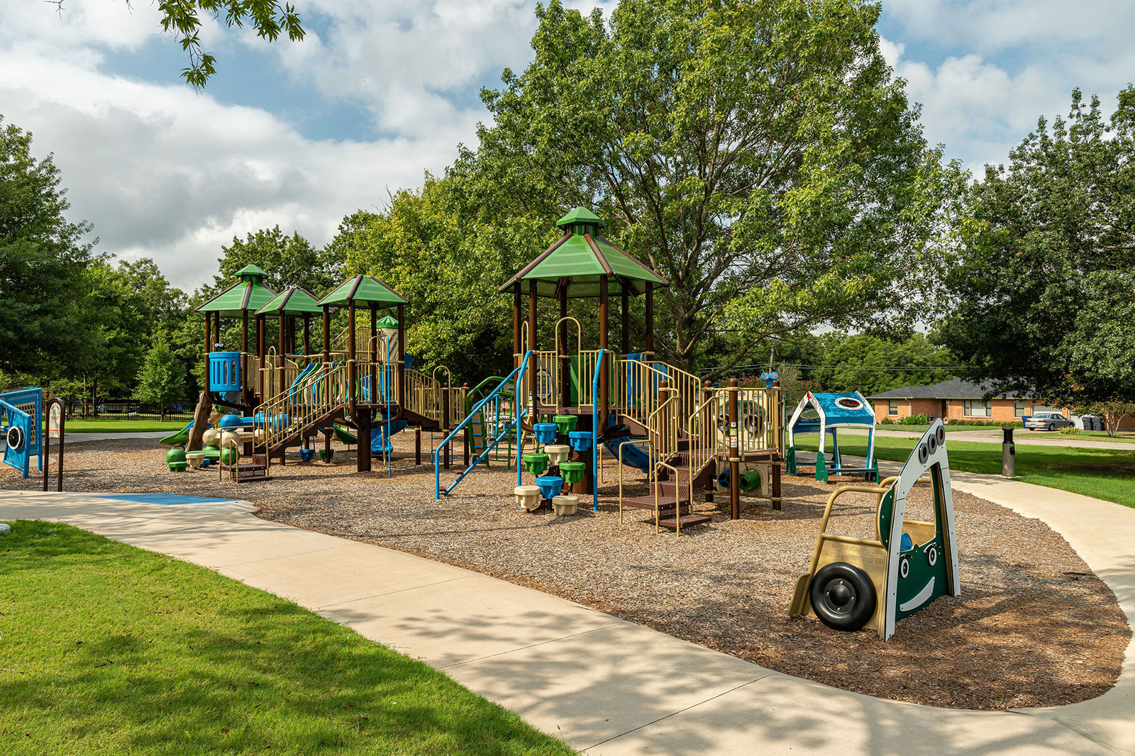 Playground with multiple climbing structures, slides, and activity panels surrounded by trees and a curved sidewalk on a sunny day.
