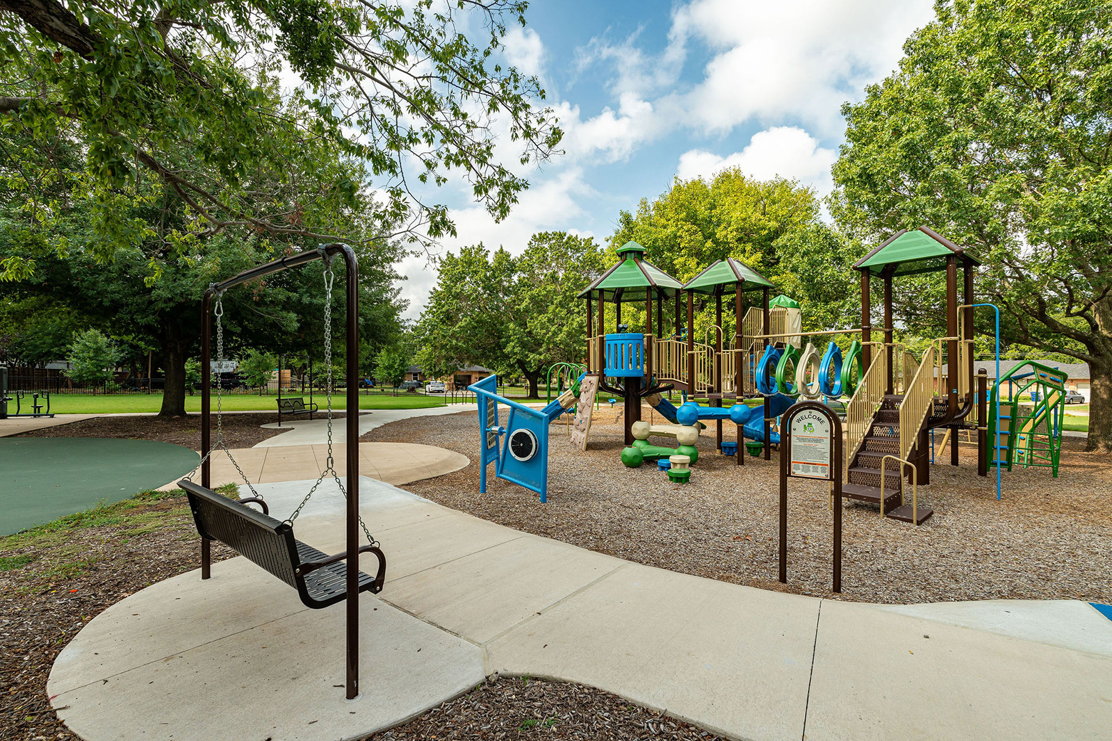 A playground with climbing structures, slides, and a swing set, surrounded by trees and a paved walking path on a sunny day.
