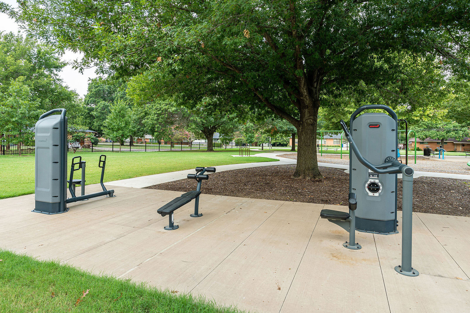 Outdoor fitness equipment is installed on a concrete pad in a park, surrounded by grass, trees, and a playground in the background.