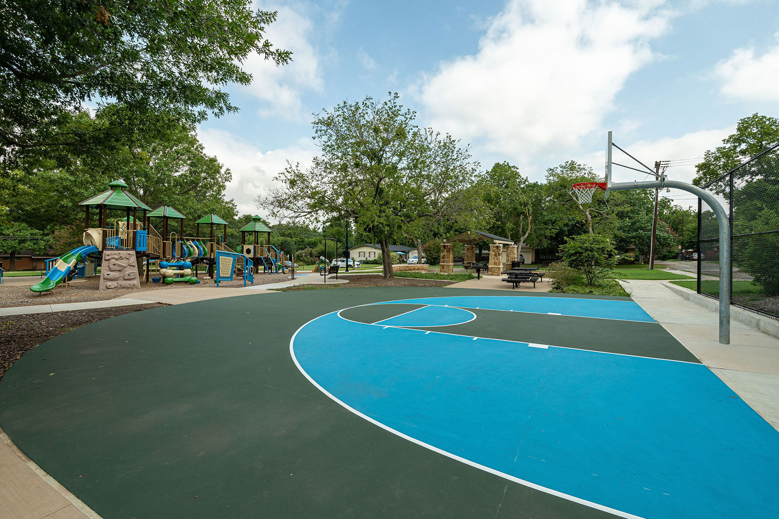 Outdoor basketball court next to a playground with slides and climbing structures, surrounded by trees and a few picnic tables on a sunny day.