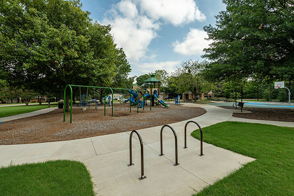 Playground with swings, slides, and climbing structures surrounded by trees, bike racks in the foreground, and a basketball court to the right.