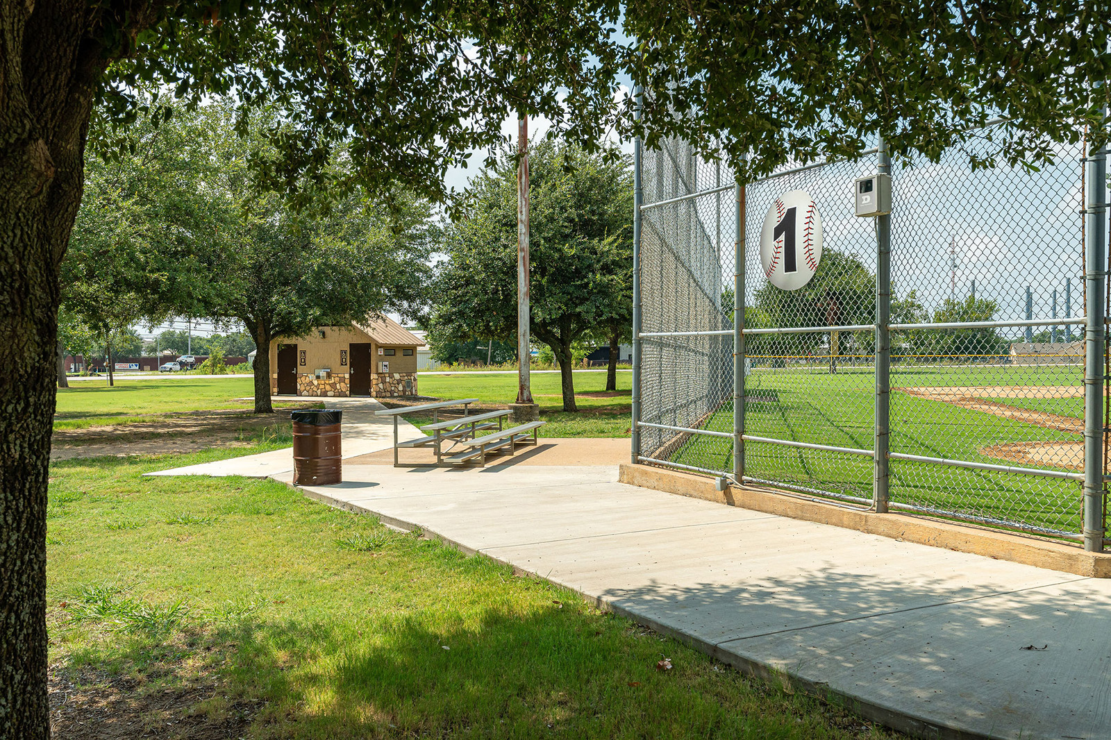 A baseball field with a chain-link fence marked "1," metal bleachers, a trash can, a small building, and trees surrounding the area on a sunny day.
