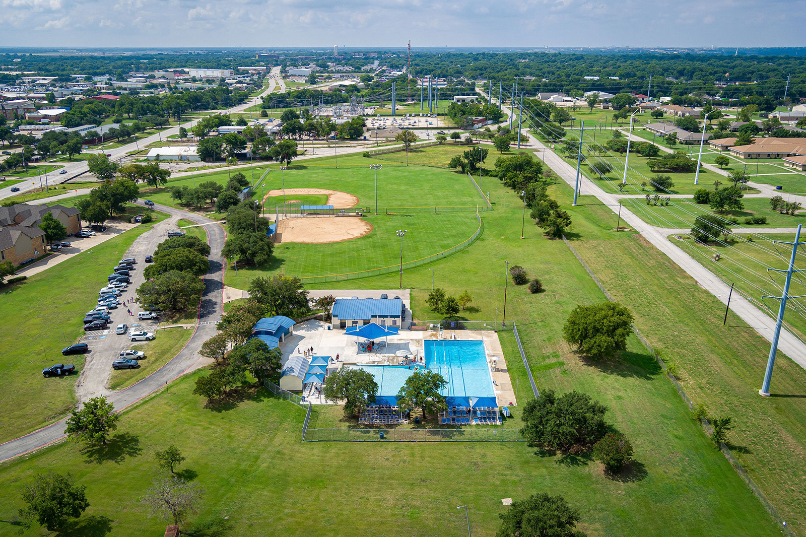 Aerial view of a community park with a baseball field, outdoor swimming pool, parking lot, and surrounding green space, bordered by roads and power lines.