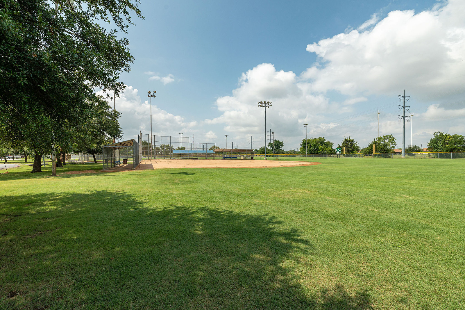 A well-maintained baseball field with green grass, a dirt infield, dugouts, light poles, and trees under a partly cloudy sky.