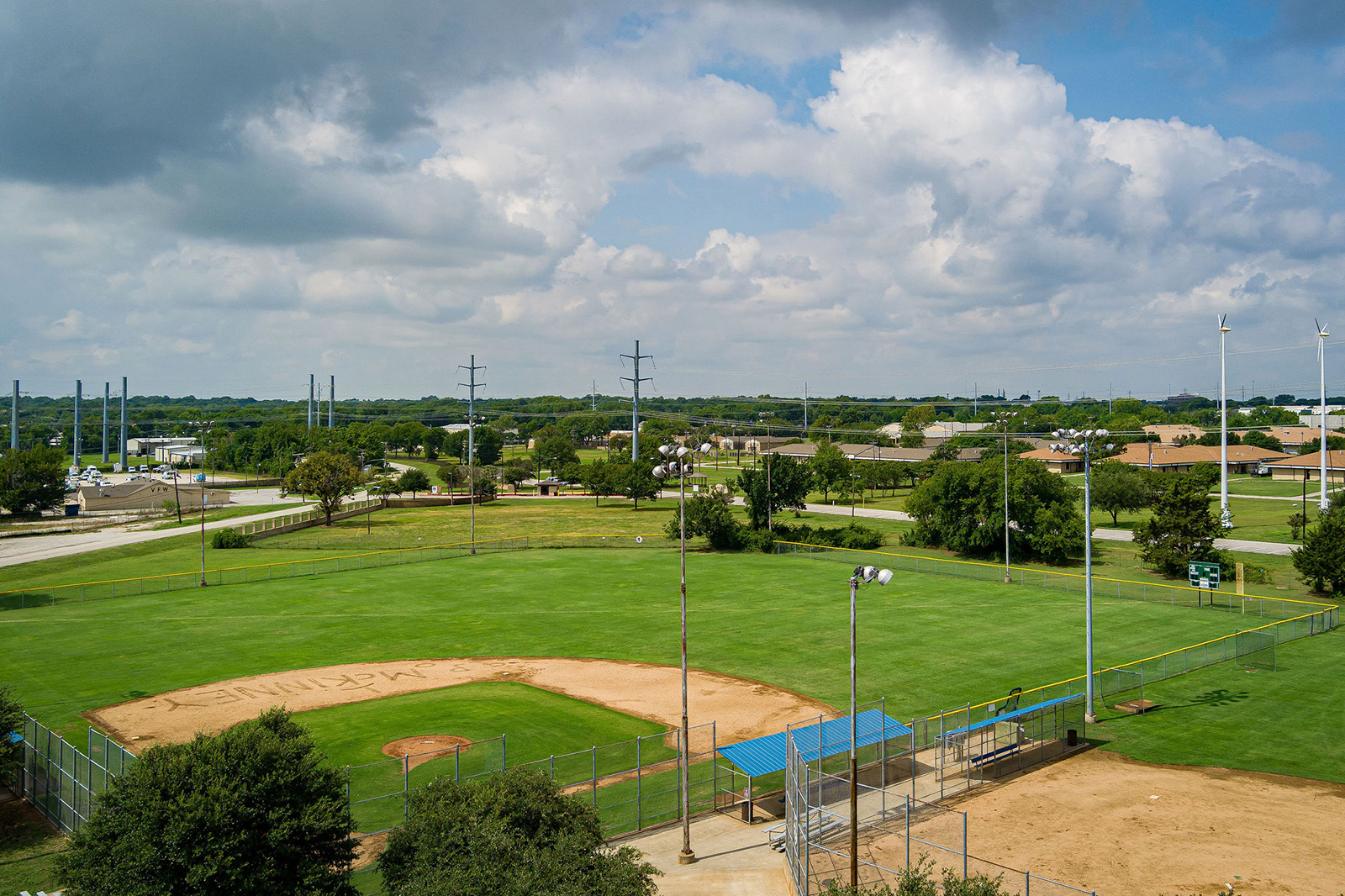 Aerial view of a baseball field with green grass, fenced perimeter, dirt infield, and surrounding suburban area under a partly cloudy sky.