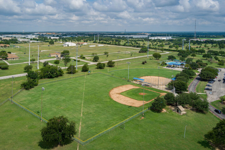 Aerial view of a baseball field surrounded by green grass, trees, parking lot, and nearby roads under a partly cloudy sky.
