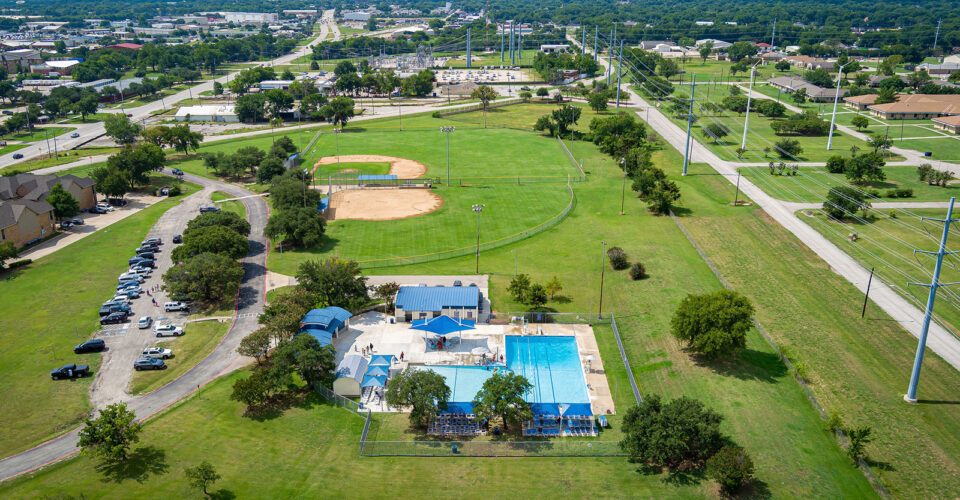 Aerial view of a public park with a swimming pool, baseball field, parking lot, green open spaces, trees, and surrounding roads in a suburban area.