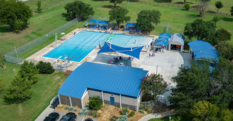 Aerial view of a public outdoor swimming pool complex with blue rooftops, shaded seating areas, nearby parked cars, and surrounding green lawns and trees.