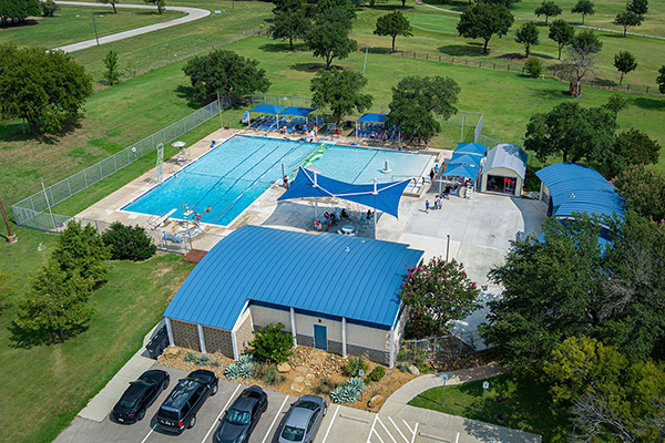 Aerial view of a public swimming pool facility with blue-roofed buildings, shaded seating areas, and people gathered around the pool, surrounded by green lawns and trees.