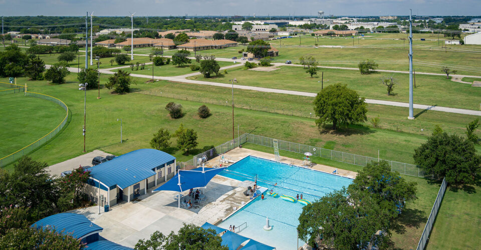 Aerial view of an outdoor swimming pool with people, surrounded by trees and open green fields, adjacent to a small building and parking area under a partly cloudy sky.