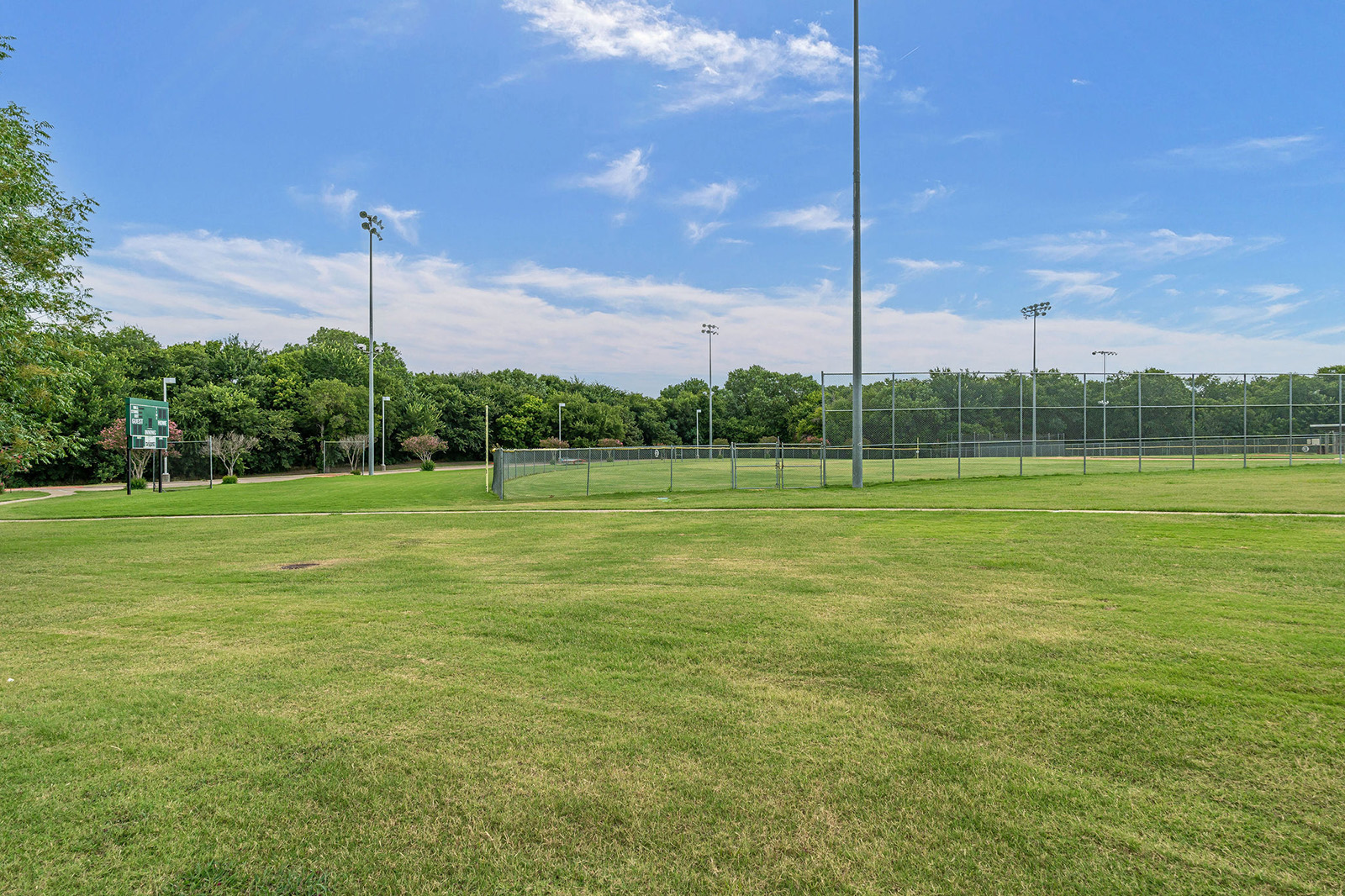 A grassy field with light poles and a fenced baseball or softball field in the background under a partly cloudy sky.