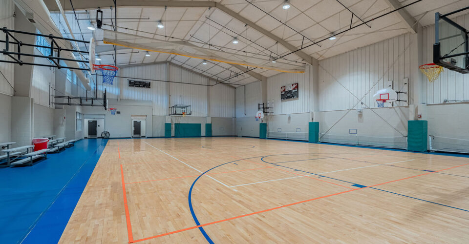 Indoor basketball court with wooden flooring, blue and orange lines, multiple hoops, bleachers on the left, and high ceiling with lights.