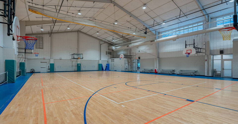 Indoor basketball gymnasium with multiple hoops, polished wooden floor, overhead lights, bleachers, and high ceiling. No people are present.