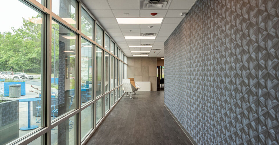 A modern hallway with dark wood flooring, patterned gray wall, large windows on the left, and white chairs at the end near a glass door.